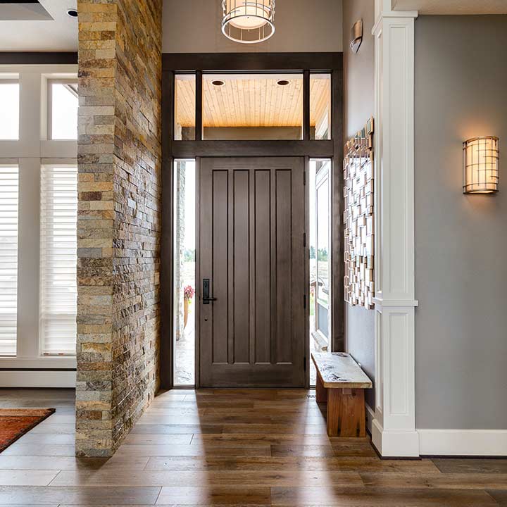 A modern entryway showcases an elegant entry door flanked by tall windows. Inside, a stone accent wall complements the wooden floors and bench. A geometric light fixture and wall sconce provide stylish lighting, creating a welcoming atmosphere.