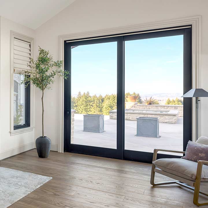 A modern living room with expansive patio doors offers a view of the spacious patio and trees outside. The room features a potted plant, an armchair, a floor lamp, and elegant wood flooring.