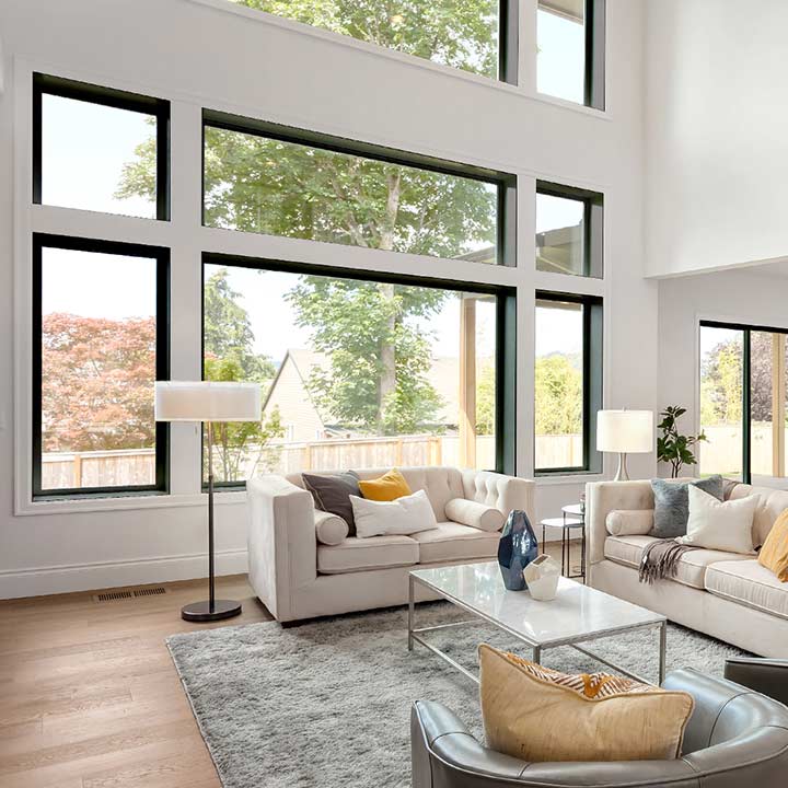 A spacious living room with large windows, two sofas, a glass coffee table, and lamps. The room has a neutral color palette with pops of yellow in the cushions. Trees are visible outside through the windows, and a textured rug covers the wood floor.
